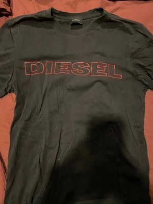 Svart t-shirt från Diesel - Snygg svart t-shirt från Diesel med stort rött logotryck på bröstet. Klassisk rund halsringning och korta ärmar. Tillverkad i mjuk bomull som känns skön mot huden. Perfekt för dig som gillar streetwear och vill ha en enkel men stilren look.
