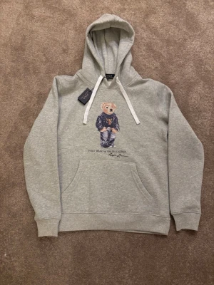 Grå hoodie Polo Bear Ralph Lauren - Grå hoodie från Ralph Lauren med Polo Bear-tryck framtill. Klassisk känguruficka, vita dragsnören och mjukt material. Perfekt för en avslappnad och trendig stil. Ikonisk björn i cool outfit på bröstet.