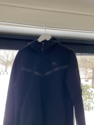 Svart Nike hoodie med dragkedja - Säljer en svart hoodie från Nike med dragkedja framtill och diskret Nike-logga på bröstet. Hoodien har huva och stilrena detaljer i form av svarta band över bröstet. Perfekt för dig som gillar sportig och clean stil.