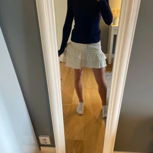 Vit volangkjol i jeans  - Supersöt vit kortkjol i jeans med flera volanger och råa, fransiga kanter. Kjolen har klassiska jeansdetaljer som fickor och bälteshällor. Perfekt för dig som vill ha en trendig och lekfull look.