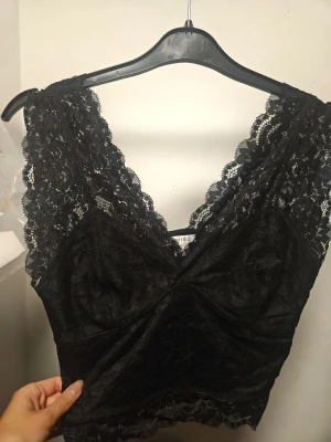 Svart spetsig linnetopp från Amisu - Supersnygg svart linnetopp från Amisu i helspets med v-ringning och breda axelband. Toppen är croppad och har fina spetsdetaljer både fram och bak. Använd nån gång och i fint skick! Bild från samma fast vit