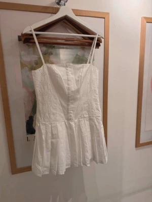 Vit kort klänning med broderi - Söt vit kortklänning med tunna axelband och broderade detaljer längs sidorna. Perfekt för studenten!