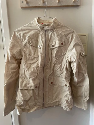 Peak Performance Safarijacka - Stilren beige vindjacka med dragkedja framtill, fyra fickor med lock och knappar samt justerbart bälte i midjan. Jackan har hög krage och långa ärmar, perfekt för vår och höst.