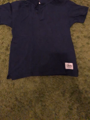 Mörkblå t-shirt från Junkyard - En stilren mörkblå t-shirt från Junkyard med kort ärm och klassisk passform. T-shirten har en liten etikett med logga nertill på framsidan och knappar vid halsen. Perfekt för en avslappnad och cool look.