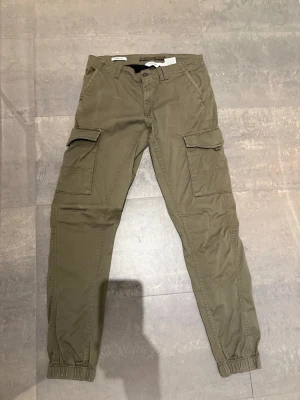 Olivgröna cargobyxor med fickor - Snygga olivgröna cargobyxor med flera stora fickor på sidorna och bak. Byxorna har resår vid bensluten och klassisk cargomodell med avslappnad passform. Perfekta för en streetwear-look och tillverkade i bomullstyg.