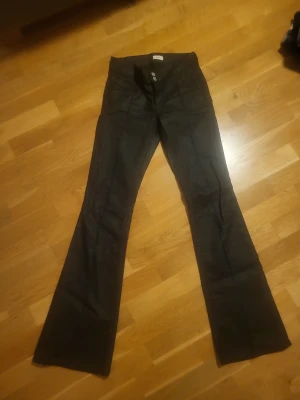 Svarta coated bootcut jeans med knappar - Säljer ett par svarta coated bootcut jeans med knäppning fram och klassiska fickor både fram och bak. Jeansen har en lätt utsvängd passform nedtill och är perfekta för dig som gillar en retrostil. Materialet känns mjukt och följsamt.