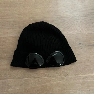 Cp company mössa - Säljer en svart ribbstickad mössa från C.P. Company med ikoniska glasögon på uppviket. Mössan är tillverkad i mjukt material och har en klassisk passform med bred kant. Perfekt för dig som vill sticka ut.