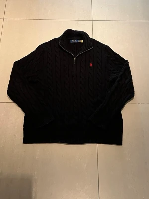Svart kabelstickad tröja Polo Ralph Lauren - Kabelstickad svart tröja från Polo Ralph Lauren med halv dragkedja och röd broderad logga på bröstet. Tröjan har hög krage och ribbade muddar. Perfekt för dig som gillar klassisk och stilren design.