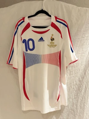 Frankrike landslags tröja Zidane #10 - Säljer en vit Frankrike fotbollströja med blått nummer 10, röda och blå detaljer samt guldiga broderier. Tröjan har korta ärmar och är tillverkad i lätt, ventilerande material. Perfekt för match eller träning.