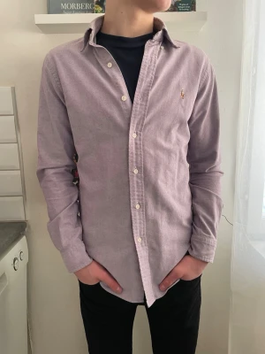 Ljuslila skjorta från Ralph Lauren - Snygg ljuslila skjorta från Ralph Lauren med klassisk krage och knappar framtill. Skjortan har lång ärm och den ikoniska broderade loggan på bröstet. Tillverkad i mjuk bomull som känns skön mot huden. Perfekt för en clean och stilren look.