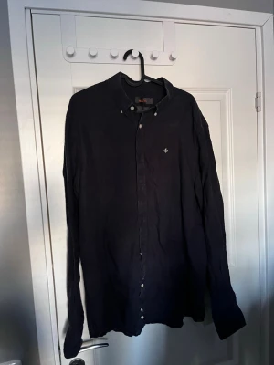 Svart skjorta från Morris - Stilren svart skjorta från Morris. Skjortan har klassisk krage med knappar, långa ärmar och diskret logga broderad på bröstet. Perfekt för en avslappnad men snygg look.