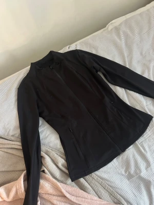 Svart träningskortet - Snygg svart träningsjacka med hel dragkedja framtill och två sidofickor med dragkedja. Figurnära passform och diskret logga på ryggen. Perfekt för cykling eller löpning under kyligare dagar.