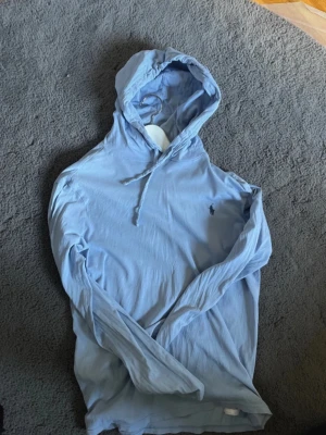 Ljusblå hoodie från Polo Ralph Lauren - Säljer en ljusblå hoodie från Polo Ralph Lauren med klassisk huva och dragsko. Hoodien har lång ärm och den ikoniska lilla loggan broderad på bröstet. Tillverkad i mjukt och tunt material, perfekt för en avslappnad stil.