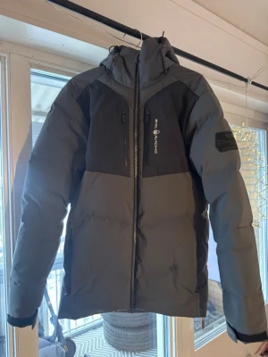 Grå dunjacka Sail Racing med huva - Säljer en grå och svart dunjacka från Sail Racing med huva och dragkedja framtill. Jackan har två bröstfickor med dragkedja, logga på bröstet och patch på ärmen. Perfekt för kalla vinterdagar och riktigt snygg streetstil.