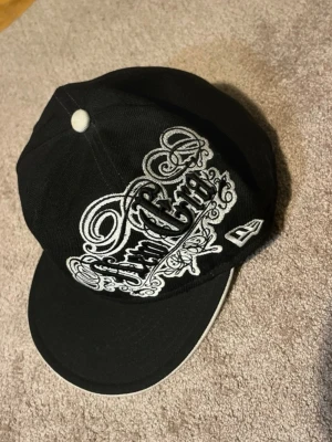 Svart New Era keps med brodyr - Säljer en svart keps från New Era med stor vit brodyr på framsidan och logga på sidan. Kepsen har platt skärm och vit detalj på toppen. Materialet är klassisk bomullscanvas och designen är streetinspirerad med tydliga kontraster.