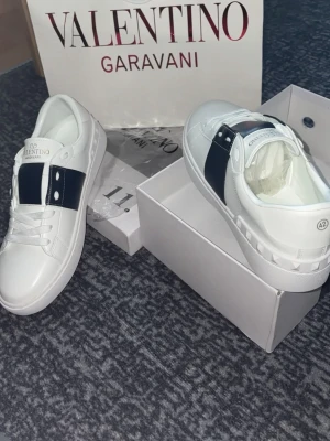 Valentino Garavani vita sneakers - Snygga vita sneakers från Valentino Garavani med en blå bred rem över ovandelen och silverfärgade nitar på sidan. Skorna är i läder och har klassisk låg profil med vita snören och diskret logga på plösen.