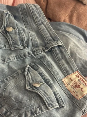 True Religion Joey ljusblå flare jeans - ALDRIG ANVÄNDA. Säljer ett par ikoniska True Religion Joey jeans i ljusblå tvätt med låg midja och flare-ben. Snygga detaljer som stora ficklock med silverknappar och klassisk logga bak. Perfekta för dig som gillar vintagevibbar och streetstyle.