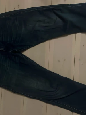 Mörkblå Replay jeans herr - Säljer ett par mörkblå jeans från Replay med snygga slitningar och klassisk femficksmodell. Jeansen har raka ben och detaljer som Replay-logga på myntfickan. Perfekta för dig som gillar stilrena och bekväma jeans.