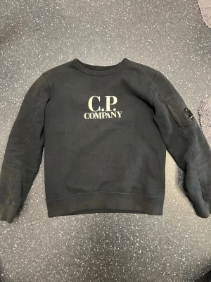Svart sweatshirt från C.P. Company - Svart sweatshirt från C.P. Company med klassisk logga på bröstet och en cool linsdetalj på ena ärmen. Tröjan har rund hals, ribbade muddar och är tillverkad i mjuk bomull. Perfekt för dig som gillar streetwear och vill ha något stilrent med en unik touch.  Priset kan diskuteras den är äkta 