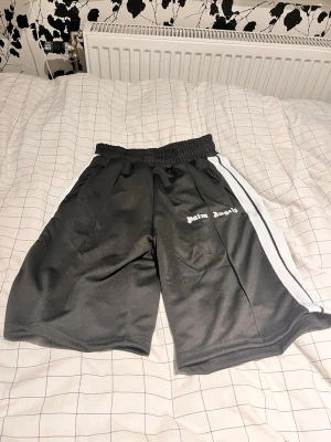 Svarta Palm Angels shorts med vit rand - Palm Angels shorts i svart med vit rand längs sidorna och logga tryckt på höger ben. De har elastisk midja och är tillverkade i ett glansigt syntetmaterial. Perfekta för en sportig och trendig look. Är 176 och de var för stora för mig