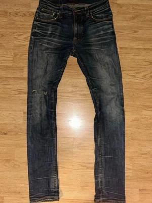 Nudie jeans  - Säljer ett par nudie  jeans i storlek w31 L34 med ett hål i knät vid högra sidan, mörkblå tvätt med snygga slitningar och whiskers. Klassisk femficksmodell med orange sömmar och låg midja. Jeansen har en smal passform och är tillverkade i slitstarkt denimtyg. Perfekta för dig som gillar en avslappnad men trendig look. HÖR AV DIG VID INTRESSE 
