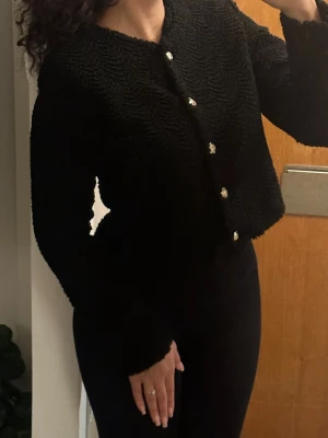 Svart cardigan med guldknappar - Stilren svart cardiganjacka med vågig struktur och dekorativa guldknappar framtill. Jackan har rund halsringning och långa ärmar, perfekt för att lyfta en enkel outfit. Materialet är mjukt och ger en lyxig känsla.