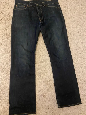 Mörkblå Levi's 513 jeans - Säljer ett par klassiska mörkblå Levi's 513 jeans i relaxed fit. Jeansen har fem fickor, gul kontrastsöm och den ikoniska läderlappen bak i midjan. Tillverkade i kraftig denim med snygg tvätt och tidlös look. Perfekta för dig som gillar stilrena och slitstarka jeans.