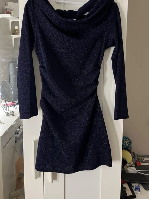 Mörkblå glittrig kortklänning H&M - Snygg mörkblå kortklänning från H&M med glittrigt tyg och långa ärmar. Klänningen har en bred, draperad urringning och figurnära passform som ger en elegant siluett. Perfekt för dig som vill sticka ut  lite extra glitter.