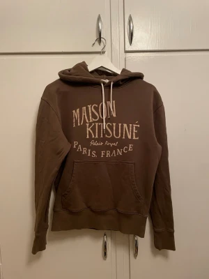  Maison Kitsuné hoodie - Snygg olivgrön (ser brun ut på bilderna) hoodie från Maison Kitsuné med stor vit logga och texten 'Palais Royal Paris, France' på bröstet. Hoodien har huva med vita dragsnören och en stor magficka. Perfekt för en avslappnad och trendig look. Effekt på bild 3! Annars bra skick