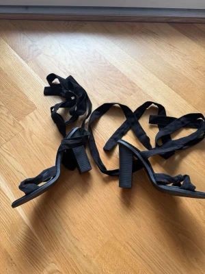 Svarta klackade sandaler Wildflower - Säljer ett par svarta sandaler från Wildflower med höga blockklackar och långa knytband som lindas runt benet. Skorna har öppen tå och är i mockaimitation, vilket ger en trendig och stilren look. Perfekta för att lyfta din outfit.