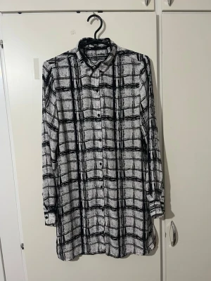 Vit och svart rutig skjorta - Långärmad skjorta i vitt med svart rutig mönster. Skjortan har klassisk krage, knäppning framtill och en loose passform. Perfekt för dig som gillar en stilren men ändå edgy look.