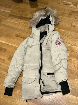 Beige parkas från Canada Goose strl M  - Snygg beige parkasjacka från Canada Goose med svart innerfoder och avtagbar huva med fuskpäls. Jackan har två stora fickor framtill, ribbade muddar och klassisk logotyp på ärmen. Perfekt för kalla dagar och riktigt varm.  OBSS! Fläckar på armarna. Inga fel på foder. Äkta !!!