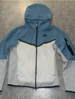 Nike Tech Fleece hoodie  - Nike Tech Fleece hoodie med dragkedja i blått, grått och vitt. Har huva, svart Nike-logga på bröstet och en praktisk ficka med dragkedja på ärmen. Snygg kontrastdesign och sportig look. Storlek XS