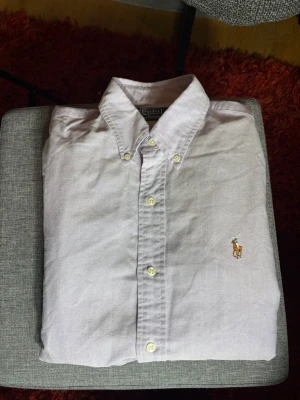 Ljuslila skjorta från Polo Ralph Lauren - Klassisk ljuslila skjorta från Polo Ralph Lauren med button-down krage och broderad logga på bröstet. Skjortan har långa ärmar, vita knappar och är tillverkad i mjuk bomull. Perfekt för en clean och stilren look.