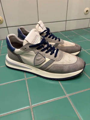 Gråa sneakers från Philippe Model - Snygga sneakers från Philippe Model i grått och vitt med blå detaljer. Ovandelen är i en mix av mocka och mesh, med blå skosnören och vit/blå sula. Klassisk låg modell med sköldlogga på sidan och vadderad häl för extra komfort.