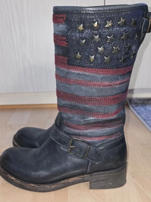 Stövlar från Scorett  - Stövlar med amerikansk flagga i rött och blått, dekorerade med stjärnor upptill. Stövlarna har spänne vid ankeln och grov sula. Dragkedja på insidan för enkel påtagning. Perfekta för dig som vill sticka ut.