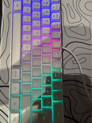 Vit mekanisk RGB-tangentbord - Säljer ett kompakt vitt mekaniskt tangentbord med RGB-belysning som lyser i flera färger. Tangentbordet har nordisk layout och ansluts med kabel. Perfekt för gaming eller studier, och ger snygg effekt på skrivbordet.