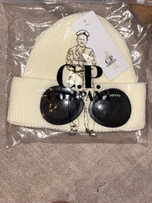 Vit mössa med goggles från C.P. Company - Snygg vit ribbstickad mössa från C.P. Company med ikoniska svarta goggles framtill. Mössan har uppvikt kant och är tillverkad i ett mjukt, stretchigt material som sitter skönt på huvudet. Perfekt accessoar för en trendig streetwear-look.