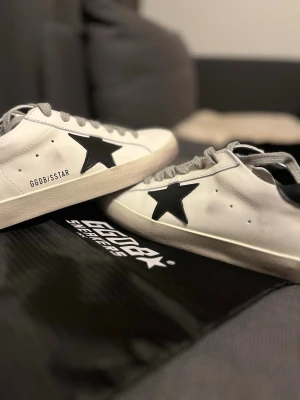 Golden Goose Svart/vita st 42 - Golden Goose sneakers i vitt skinn med svart stjärna på sidan och svart hälparti. Grå snörning och klassisk platt sula. Snygg kontrast mellan färgerna och ikonisk branding på hälen. Perfekta för dig som gillar streetstyle och vill sticka ut.