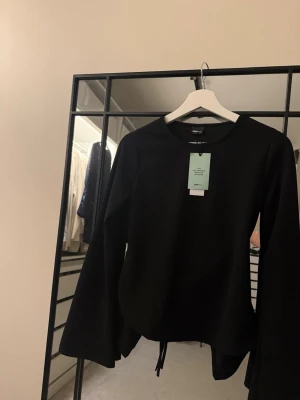 Svart långärmad topp med knyt från Gina Tricot - Säljer en svart långärmad topp från Gina Tricot med rund hals och snygga utsvängda ärmar. Toppen har knyt i ryggen för en cool detalj och är gjord av 97% återvunnen polyester. Perfekt att styla med jeans eller kjol för en trendig look.