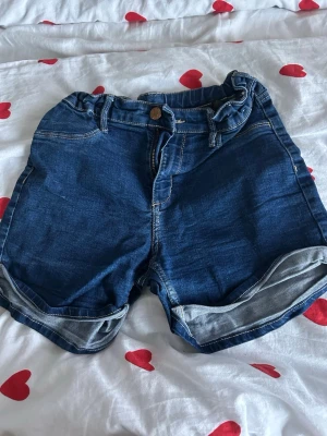 Blå denimshorts från &Denim - Säljer ett par klassiska blå jeansshorts från &Denim i storlek 152. De har normal passform, uppvikta benslut, två fickor bak och fram samt knapp och dragkedja. Perfekta för varma dagar och har en snygg, enkel look.