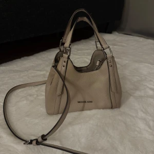 Beige handväska från Michael Kors - Snygg ägg vit handväska från Michael Kors i mjukt skinn med silverfärgade metalldetaljer. Väskan har både handtag och en justerbar axelrem, samt en diskret logga framtill. Perfekt storlek för det viktigaste och stilren design som passar till många outfits.