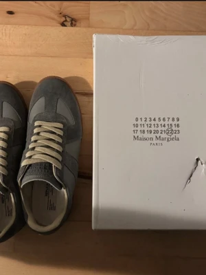 Maison Margiela grå sneakers - Maison Margiela sneakers i grått med beige snörning och gummisula. Skorna har en klassisk rund tå och är tillverkade i en mix av mocka och skinn. Snygg retrostil med diskreta paneler och branding på sulan. Perfekta för dig som gillar stilrena och exklusiva sneakers.