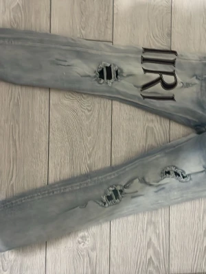 Ljusblå slitna jeans med Amiri tryck - Unika ljusblå jeans med slitningar och hål på benen samt stora svarta bokstavstryck på både fram- och baksida. Jeansen har en rak passform och klassiska bakfickor. Perfekta för dig som vill sticka ut med en streetwear-look.