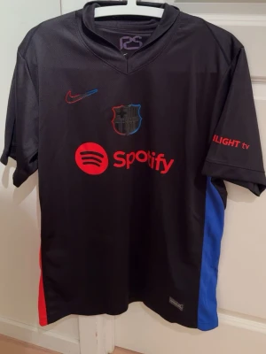 Svart FC Barcelona Nike t shirt - Säljer en svart FC Barcelona t shirt från Nike med röda och blå detaljer på sidorna. Tröjan har Dri-Fit material, Spotify-logga på bröstet och klubbmärke i mitten. Snygga röda tryck på ärm och rygg, perfekt för dig som älskar Barca. Den är i storlek L men skulle även passa M då den är lite liten på mig