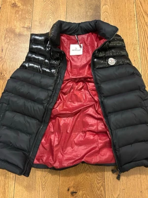 Svart dunväst från Moncler - Säljer en svart dunväst från Moncler med glansiga paneler upptill och klassisk Moncler-logga på bröstet. Insidan är röd och västen har en hög krage samt dragkedja framtill. 
