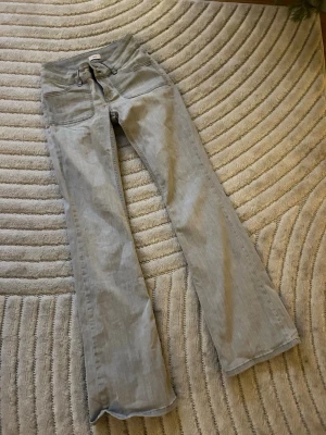 Grå bootcut jeans från Nelly - Säljer ett par gråa bootcut jeans från Nelly med låg midja. Jeansen har en snygg, lätt utsvängd passform och är tillverkade i mjukt bomullsmaterial med lite stretch. Perfekta för dig som gillar en retro vibe och vill ha ett par jeans som sticker ut. Lite använda, därav lite slitage längst ner vid fötterna baktill.