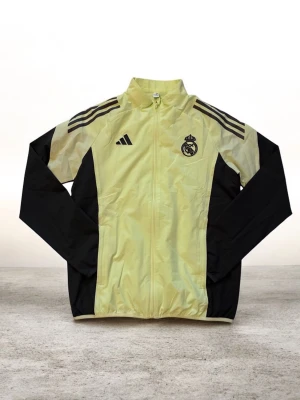 Real Madrid vindjacka från Adidas - Gul och svart vindjacka från Adidas med Real Madrid-logga på bröstet och klassiska Adidas-ränder på axlarna. Jackan har hel dragkedja framtill, ståkrage och långärmade svarta ärmar. Perfekt för dig som vill ha sportig stil och visa ditt lagstöd.