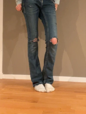 Grace in LA blå bootcut jeans med broderi - Säljer ett par blå jeans från Grace in LA med bootcut-snitt och låg midja. Jeansen har  detaljer och hål vid knäna, samt snygga broderade fickor med pärlor och mönster i guld, vitt och blått. Säljer för jag har tröttnat på dem. Jag är 175 och sitter bra i längden men sitter nog bättre om man är lite kortare, dom ser inte så låga midjade ut men dom är det