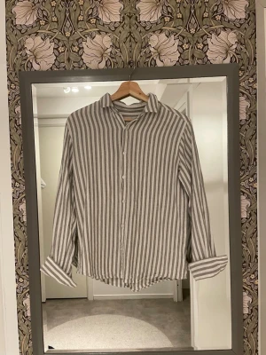 Randig skjorta från 157 i linne - Snygg skjorta från 157 i storlek S, med vertikala grå och vita ränder. Tillverkad i linne, vilket gör den luftig och skön. Klassisk krage, knappar framtill och långa ärmar som kan rullas upp för en chill look. Använd 1 gång!